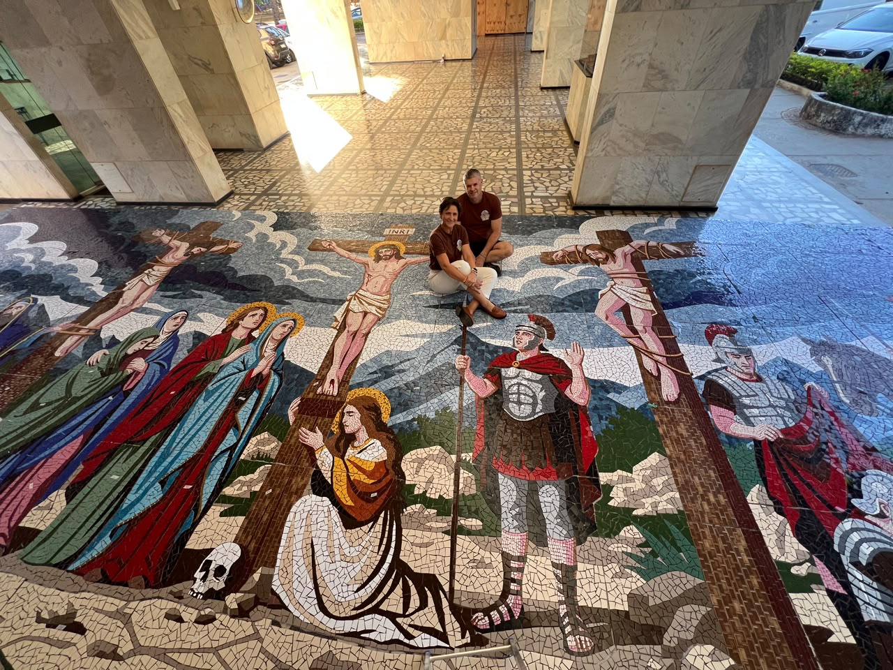 Marta e o marido Fernando sentados ao lado do mosaico sacro gigante. - Foto: reprodução/ Instagram/ @martamosaicos